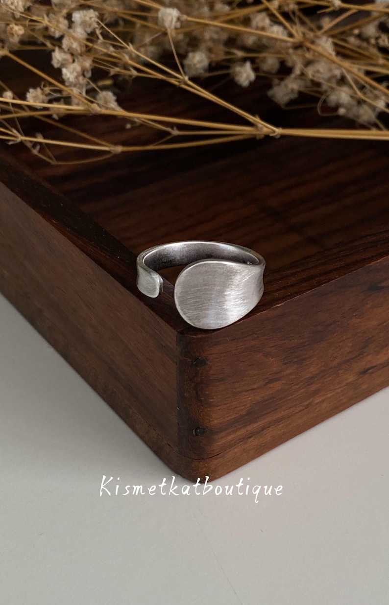 May include: A silver-toned open ring featuring a smooth, rounded, flat circular focal point with a brushed metal finish. The band is adjustable and displays a subtle, matte texture throughout. The text Kismetkatboutique is visible below.