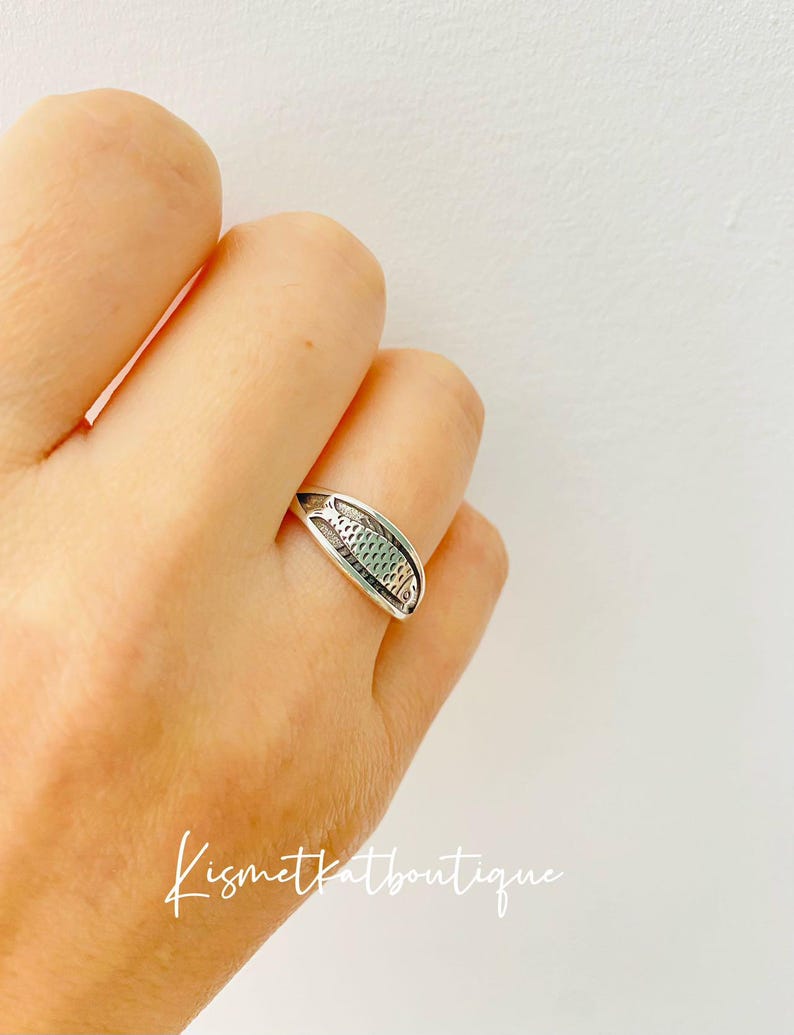May include: A silver ring with a detailed fish design. The ring is worn on a person's finger.