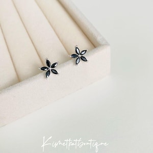 May include: A pair of silver stud earrings with black flower-shaped accents. The earrings are displayed on a white velvet background.