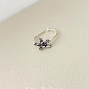 Puede incluir: Anillo ajustable de plata con un diseño de estrella de mar. La estrella de mar está texturizada con pequeños puntos.