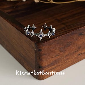 May include: A silver-toned open ring featuring a series of four-pointed star shapes along the band. The star motifs are accented with small, dark blue circular stones set within the metal. The text Kismetkatboutique is visible below the ring.