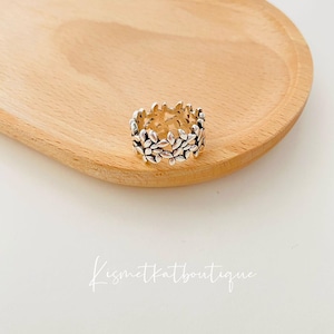 May include: A silver ring featuring a delicate design of small leaves. The ring is displayed on a wooden surface.