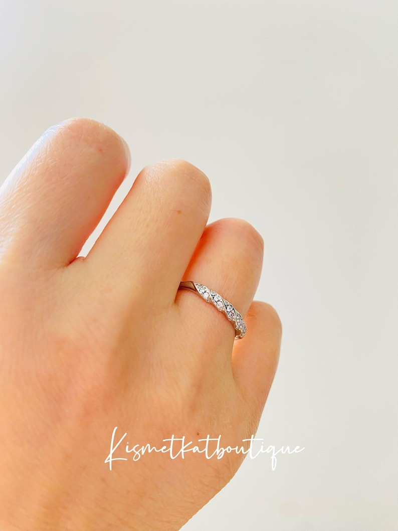 Op de afbeelding: Een zilveren ring met een rij kleine, heldere edelstenen in een herhalend patroon. De ring zit om een vinger, met de tekst "Kismetkatboutique" in cursief.