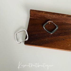 Puede incluir: Dos pendientes de aro cuadrados de plata en un expositor de madera. Los pendientes son de diseño simple y moderno.