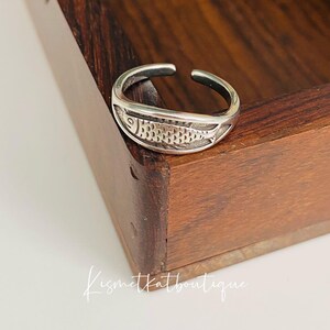 May include: A silver adjustable ring with a detailed fish design. The ring is sitting on a wooden surface.