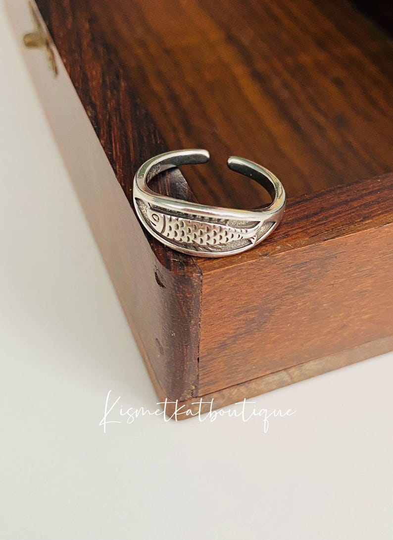 May include: A silver adjustable ring with a detailed fish design. The ring is sitting on a wooden surface.