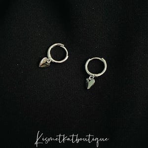 May include: A pair of silver hoop earrings with a small heart charm hanging from each hoop.