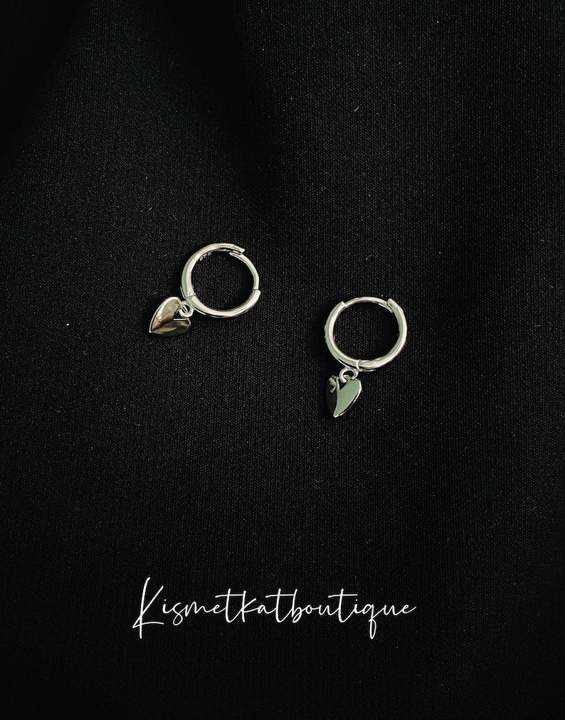 May include: A pair of silver hoop earrings with a small heart charm hanging from each hoop.