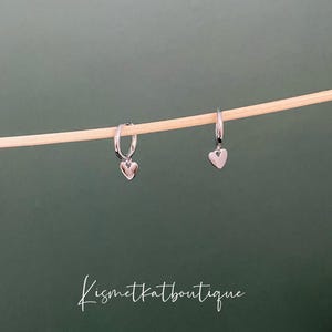 May include: A pair of silver hoop earrings with a small heart charm hanging from each hoop.