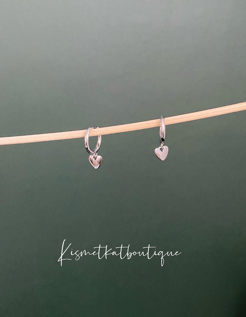 May include: A pair of silver hoop earrings with a small heart charm hanging from each hoop.