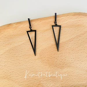 May include: A pair of black triangle earrings. Each earring has a small rectangular post and a hollow, open triangle shape. The earrings are presented on a wooden surface. The text "kismetkatboutique" is visible.