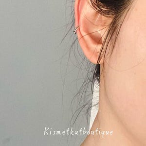 May include: A close-up of an ear with a small, silver ear cuff. The ear cuff has a geometric design. The text "Kismetkatboutique" is written in cursive at the bottom of the image. The background is a solid grey.