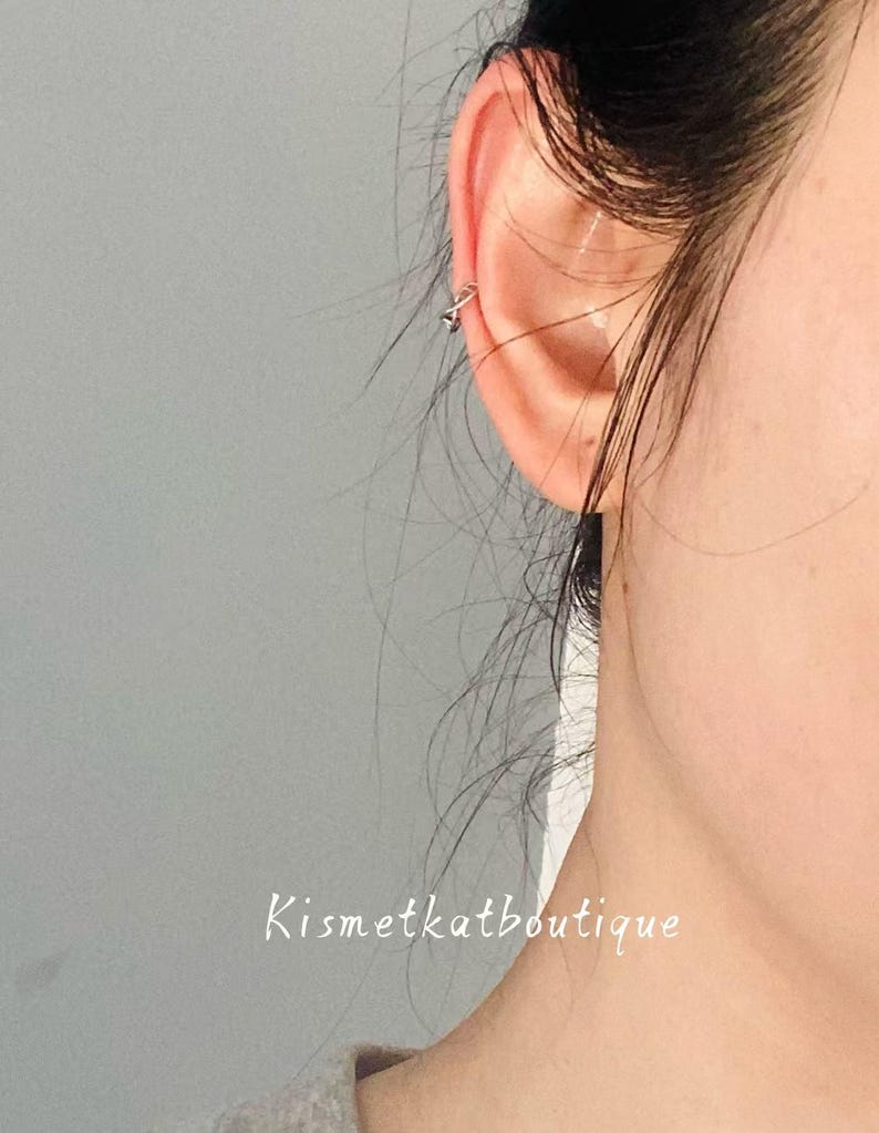 May include: A close-up of an ear with a small, silver ear cuff. The ear cuff has a geometric design. The text "Kismetkatboutique" is written in cursive at the bottom of the image. The background is a solid grey.