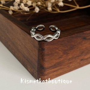 May include: A silver-toned open ring featuring a continuous twisted rope or infinity-style pattern. The band has a polished metallic finish and an adjustable gap at the top. The text Kismetkatboutique is visible in white lettering below the ring.