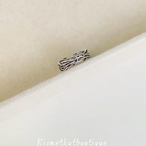 以下が含まれることがあります： 織り模様のシルバーカラーのリング。リングは、複数の金属ストランドが絡み合って作られており、テクスチャード加工されたバンドスタイルのリングになっています。リングは白い表面に表示されています。
