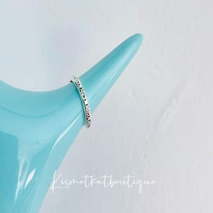 May include: A silver ring with a decorative pattern on a light blue surface. The ring is on a display stand.