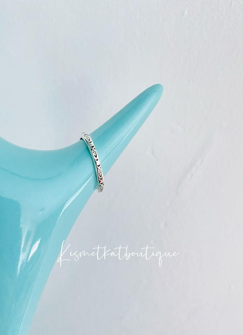 May include: A silver ring with a decorative pattern on a light blue surface. The ring is on a display stand.