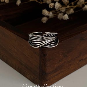 May include: A silver-colored ring with a woven design. The ring features multiple bands of metal that intertwine to create a textured, layered effect. The ring is set against a dark wooden background with dried flowers.