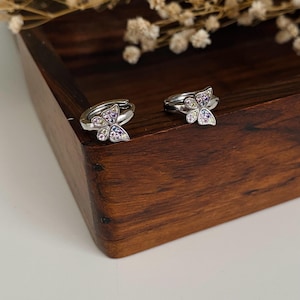 Puede incluir: Un par de pendientes de aro plateados. Cada pendiente presenta un diseño de mariposa adornado con pequeñas piedras preciosas de colores. Los pendientes se exhiben sobre una superficie de madera, con flores secas en el fondo. El texto "Kismetkatboutique" es visible.