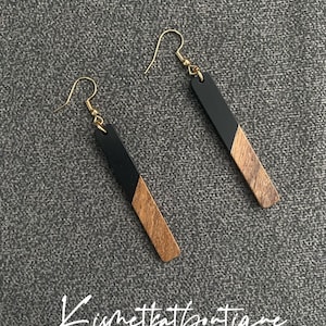 Puede incluir: Un par de pendientes de madera negra y marrón con ganchos dorados. Los pendientes son de forma rectangular y tienen una línea diagonal que separa las secciones negra y marrón.