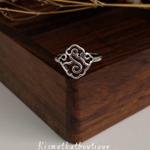May include: A silver-toned ring featuring a central openwork design shaped like a stylised letter S surrounded by cloud-like scroll patterns. The metal has a polished finish with darkened recessed areas to highlight the intricate swirling details.
