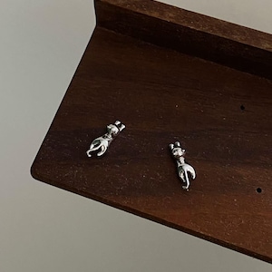 Op de afbeelding: Twee zilveren katvormige oorstekers op een donkerhouten oppervlak. De oorbellen hebben gedetailleerde kenmerken en een minimalistisch ontwerp. De tekst "Kismetkatboutique" is zichtbaar in de afbeelding.