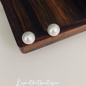 May include: Two white pearl earrings on a wooden surface. The earrings are round and have a smooth finish.