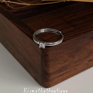 May include: A silver-toned metal ring featuring a single clear, round-cut stone set in a four-prong setting. The band is open at the back, allowing for adjustable sizing. The text Kismetkatboutique is printed in white below the ring.