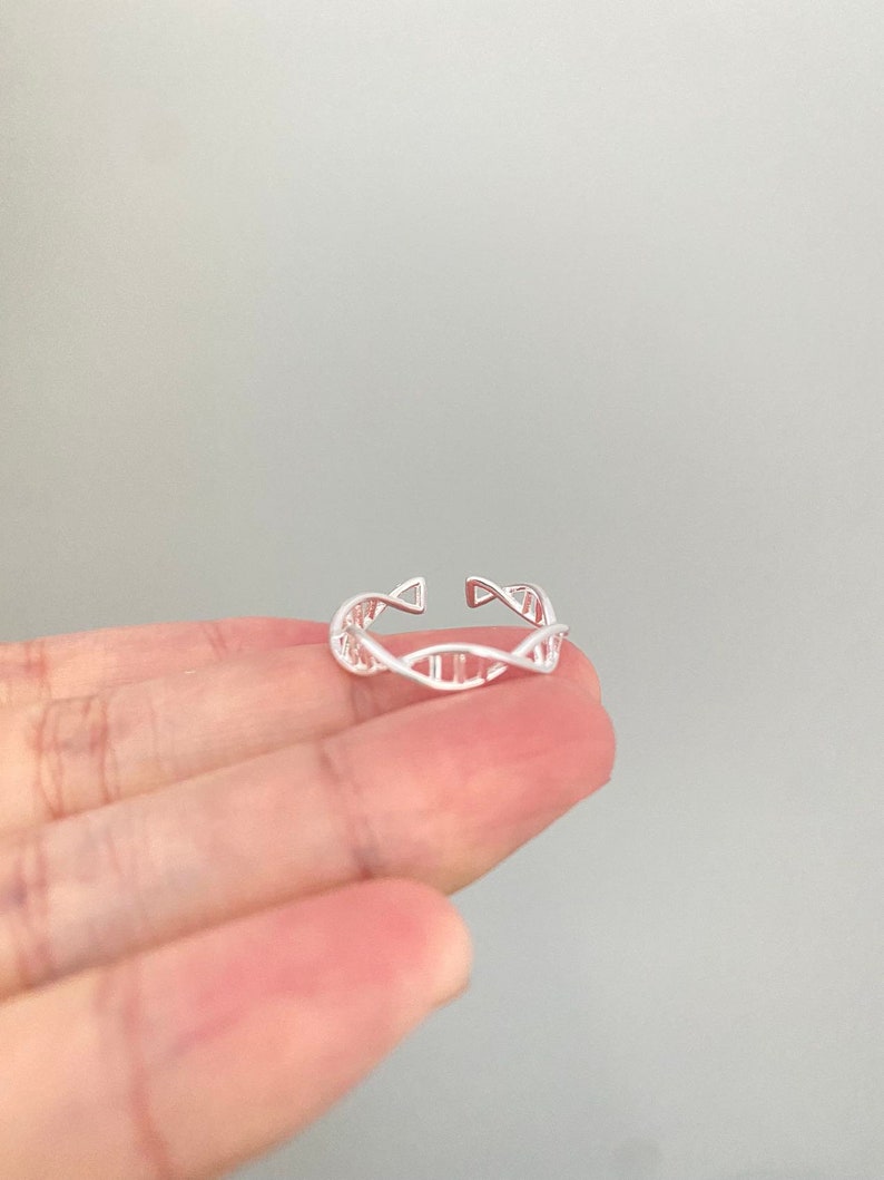 May include: A silver ring featuring a DNA double helix design. The open-ended ring is adjustable and has a detailed, openwork pattern. The ring is displayed against a neutral background.
