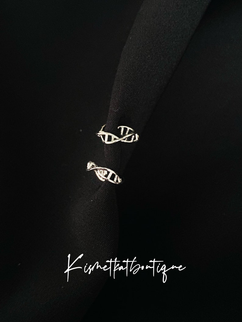 May include: Two silver DNA helix ear cuffs are shown against a black backdrop. The ear cuffs have a delicate, open design. The image also includes the text "Kismetpatboutique" in cursive script. These ear cuffs are a stylish accessory.