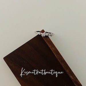 May include: A silver ring featuring a cluster of small, clear stones set in a delicate, curved design. The ring is displayed on a dark brown wooden surface with the text "Kismetkatboutique" written in white.