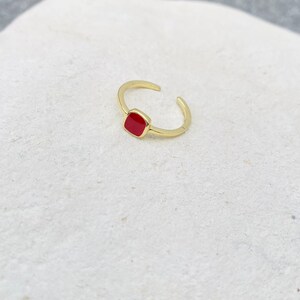 May include: A gold-colored adjustable ring with a square, red enamel center. The ring is open at the bottom, allowing for size adjustment. The ring is set against a white background.