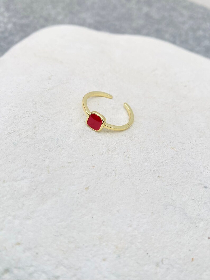 May include: A gold-colored adjustable ring with a square, red enamel center. The ring is open at the bottom, allowing for size adjustment. The ring is set against a white background.
