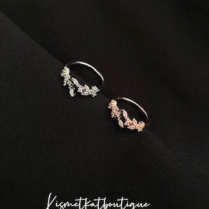 May include: Two rings, one silver and one rose gold, each featuring a leaf-shaped arrangement of clear crystals. The rings are displayed on a dark backdrop, with the cursive text "Kismetkatboutique" visible.