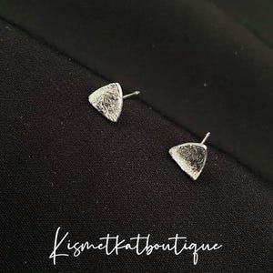 May include: A pair of silver-colored triangle stud earrings with a textured surface. The earrings are displayed on a black fabric background. The text "Kismetkatboutique" is written in cursive at the bottom of the image.