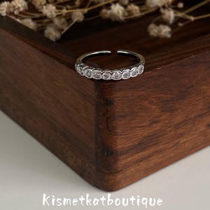 May include: A silver-toned open band ring featuring a row of small, clear, round-cut stones set in a bezel style. The ring is positioned on the corner of a dark brown wooden box, with the text Kismetkatboutique printed in white below.