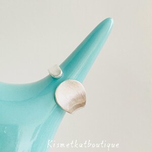 May include: A silver ring with an open band design featuring two distinct, brushed-finish circular elements of different sizes. The ring is displayed on a smooth, light blue, tapered object. The text Kismetkatboutique is visible in white.