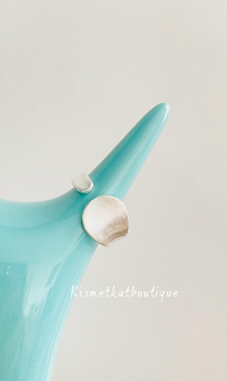 May include: A silver ring with an open band design featuring two distinct, brushed-finish circular elements of different sizes. The ring is displayed on a smooth, light blue, tapered object. The text Kismetkatboutique is visible in white.