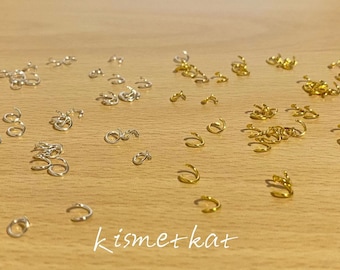 May include: A close-up image of a variety of gold and silver jump rings. The jump rings are scattered on a wooden surface. The image is labeled "kismetkat".