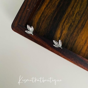 May include: A pair of silver leaf earrings on a wooden surface. The earrings are small and delicate, with a simple design. The text "kismetkatboutique" is visible in the bottom right corner of the image.