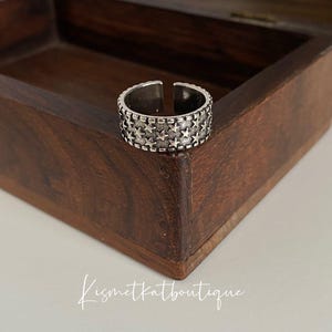 May include: A silver ring with a textured design featuring multiple rows of small stars. The ring is displayed in a wooden box.