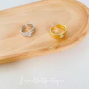 May include: Two adjustable rings with a single clear rhinestone on a wooden surface. The ring on the left is silver and the ring on the right is gold.