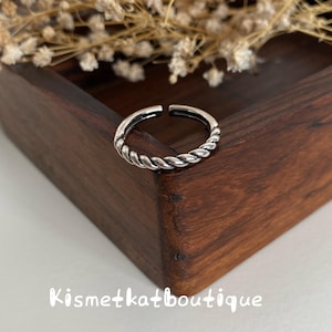 May include: A silver-toned open ring featuring a twisted rope-like texture along the band. The ring is positioned on a dark wooden surface with the text Kismetkatboutique visible in white lettering at the bottom of the image frame.