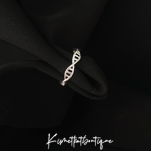 May include: A silver ring, designed as a DNA double helix, set against a black backdrop. The image includes the cursive text "Kismetkatboutique" at the bottom.