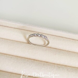 Op de afbeelding: Een zilveren ring met een gevlochten ontwerp, bezet met kleine, heldere edelstenen. De ring wordt gepresenteerd op een beige oppervlak. De tekst "Kismetkatboutique" staat in cursief.