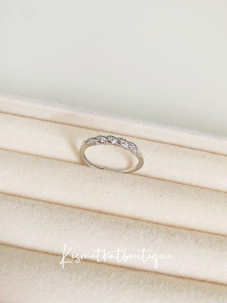 Op de afbeelding: Een zilveren ring met een gevlochten ontwerp, bezet met kleine, heldere edelstenen. De ring wordt gepresenteerd op een beige oppervlak. De tekst "Kismetkatboutique" staat in cursief.