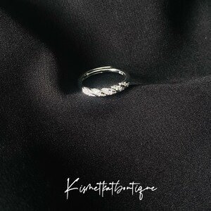 Op de afbeelding: Een zilveren ring met een getextureerde band en kleine, sprankelende versieringen. De ring is geplaatst tegen een donkere, getextureerde achtergrond. De ring is een sieraad.