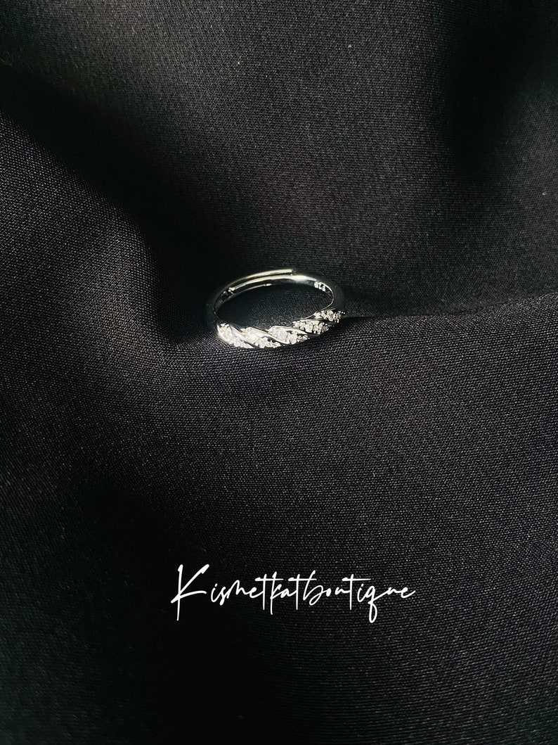 Op de afbeelding: Een zilveren ring met een getextureerde band en kleine, sprankelende versieringen. De ring is geplaatst tegen een donkere, getextureerde achtergrond. De ring is een sieraad.