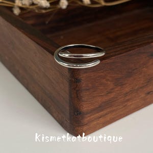 Könnte beinhalten: Ein silberfarbener Bypass-Ring mit glatter, polierter Oberfläche und einem offenen, überlappenden Design. Der Ring ist auf der Ecke einer dunkelbraunen Holzschachtel platziert. Der Text Kismetkatboutique ist in Weiß unter der Holzschachtel gedruckt.