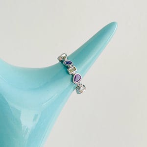 Puede incluir: Un anillo de plata con una serie de piedras preciosas de colores en varias formas, incluyendo esmeralda, pera y ovalada. Las piedras preciosas son en tonos verdes y morados. El anillo se muestra sobre una superficie azul claro. El texto "Kismetkatboutique" es visible.